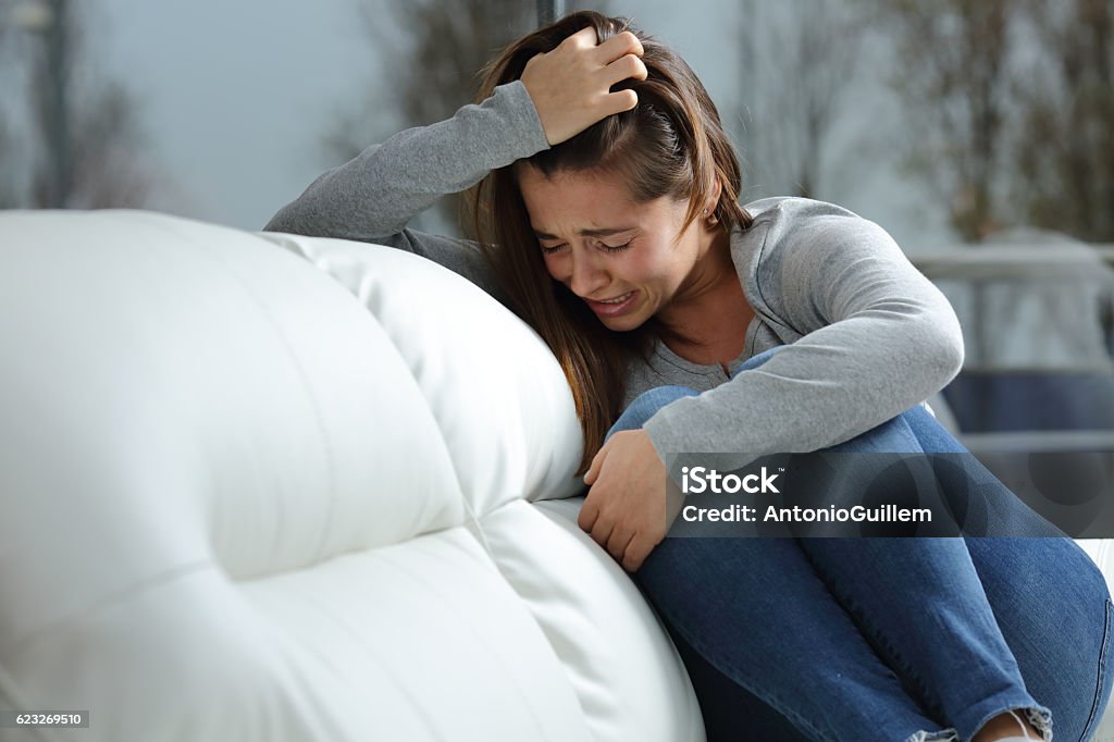 Sad girl crying alone at home Sad girl crying desperately alone sitting on a couch at home in a dark winter day Crying Stock Photo