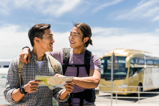A photo of hikers talking while reading map. Smiling male friends are walking by bus against sky. Backpackers are wearing casuals.
