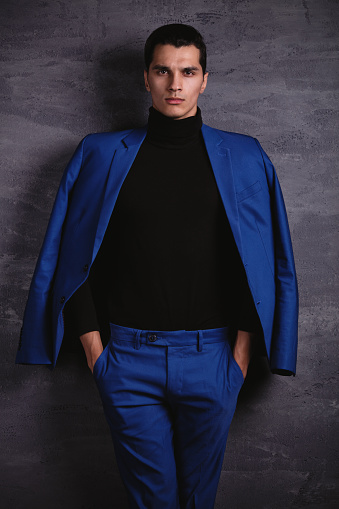 Handsome man fashion model posing a front of gray wall dressed in elegant blue jacket and pants.