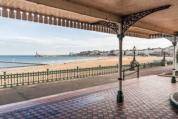 Margate Bay from a victorian shelter in Kent, South-east England, UK. It is an arching bay with a coast path running next to the shoreline. Part of margate town and harbour is visible in the distance. Victorian railings also line the beach front.