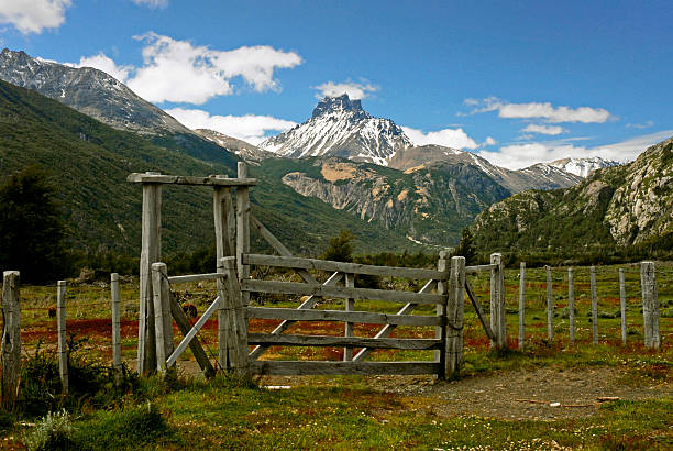 A rust-colored metal gate stands in the foreground, slightly bowed, locked but imperfectly closed.Beyond it, an open field extends toward the dark ridges under a restless sky — clouds swelling and parting in blue-white tumult.The grass below glows with faint traces of autumn bronze and late-season green, merging with the earth in subdued light.This image embodies threshold energy — a visual and emotional pause between passages.It reads simultaneously as invitation and restraint: the open road that stops short, the horizon that both beckons and refuses.In tone, it resonates as an echo of longing — the sense that beauty often resides just beyond reach, across the symbolic gate of circumstance and time.