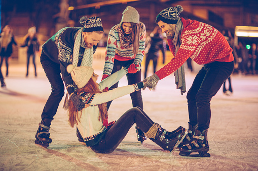 Friends having so much fun while ice skating.  Wearing warm clothing. City is decorated with christmas lights.