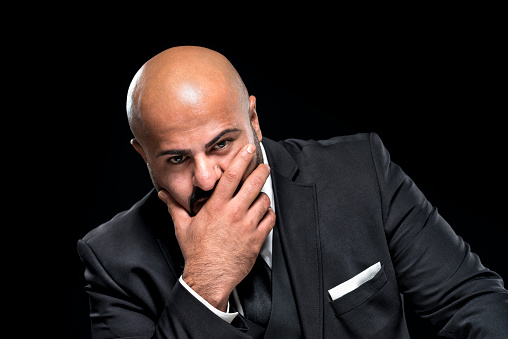 Bald businessman with black beard looking embarrassed at camera. He is wearing an elegant black suit, a white button-down shirt and a tie. Studio shot in front of a black background.