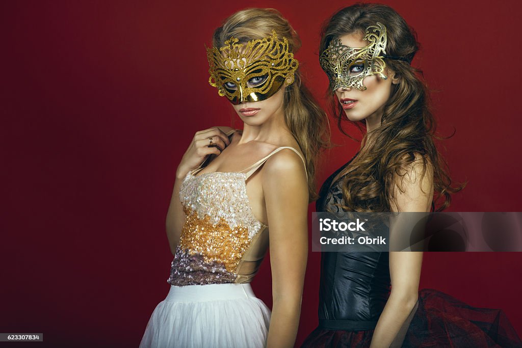 Two glam women in masks on red background Two glam gorgeous women, blonde and brunette, in golden and bronze masks wearing evening gowns standing on red background gazing intently with expressive eyes. Studio shot. Copy-space Evening Ball Stock Photo
