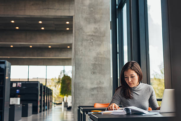 Female student doing assignments in library Indoor shot of young female student doing assignments in library. Asian woman reading textbook while sitting at college library. asian adult student stock pictures, royalty-free photos & images