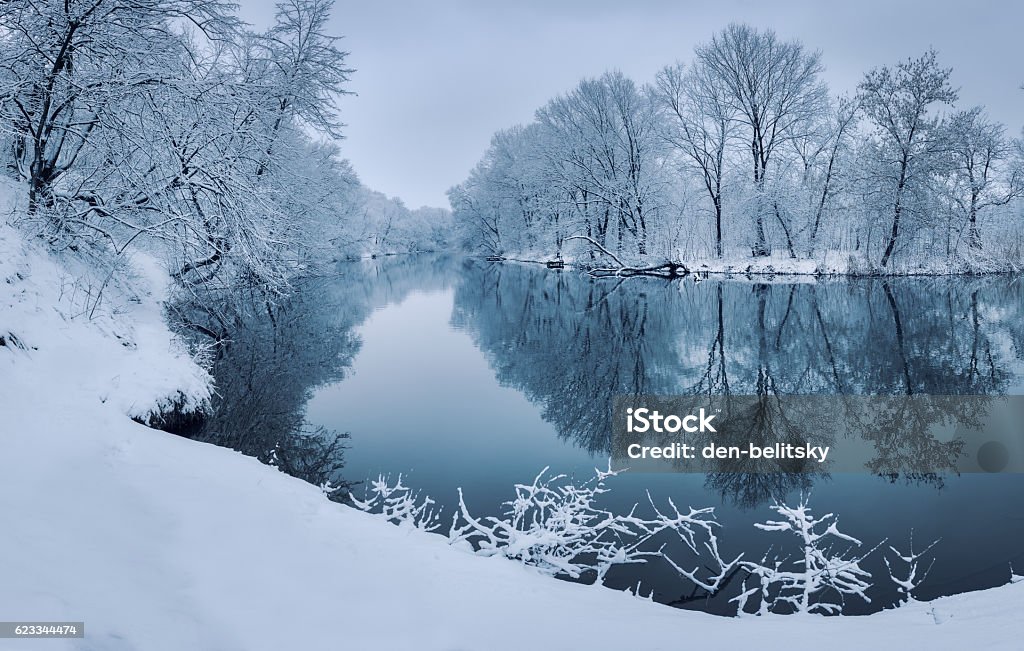 Colorful landscape with snowy trees, beautiful frozen river at sunset Winter forest on the river at sunset. Colorful landscape with snowy trees, beautiful frozen river with reflection in water. Seasonal. Winter trees, lake and blue sky. Frosty snowy river. Weather Winter Stock Photo