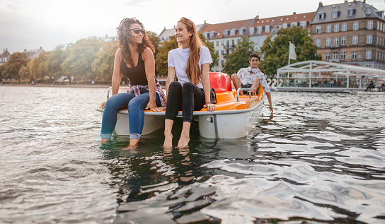Outdoors shot of young female friends sitting in front pedal boat and putting their feet in water with man in background. Teenage friends enjoying boating in the lake.