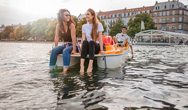 Teenage friends enjoying boating in the lake Outdoors shot of young female friends sitting in front pedal boat and putting their feet in water with man in background. Teenage friends enjoying boating in the lake. paddleboat stock pictures, royalty-free photos & images
