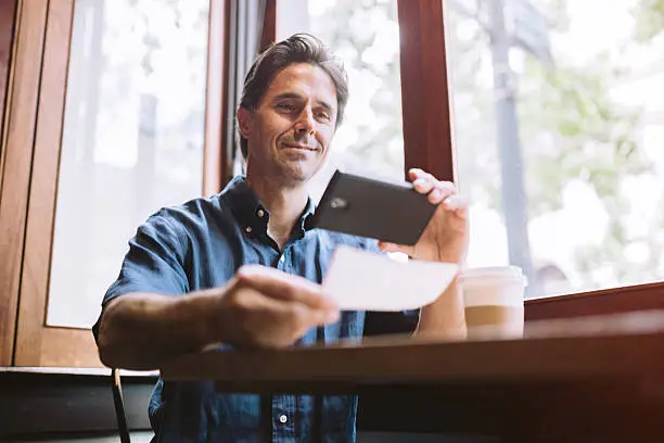 A mature smiling man in his mid 40's takes a picture with his smart phone of a check or paycheck for digital electronic depositing, also known as "Remote Deposit Capture".  He sits with a cup of coffee at a cafe.  Horizontal image with copy space.