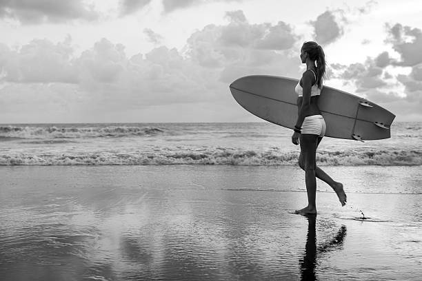 A young athletic Caucasian woman walks on the beach at low tide with her surfboard to face the ocean. The sun slowly sets down in the horizon. In Black and White mode with copy space on the left side of the image.