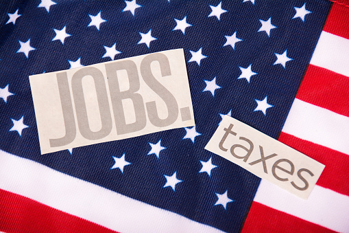 The words jobs and taxes cut out of a newspaper laying on the American flag. Importance of government in creating jobs and collecting taxes, Political picture of jobs and taxes. American flag with red, white and blue and paper cut outs.