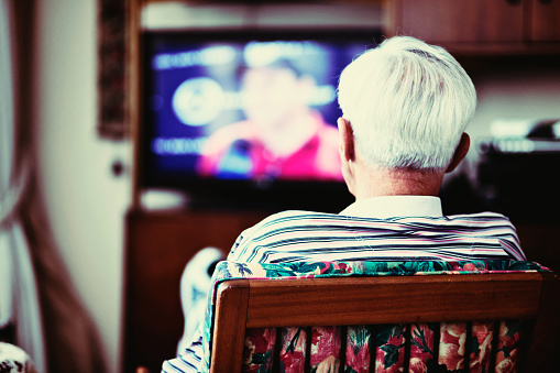 A 90-year-old old man is seen backview, seated in front of a television, alone. He may be a resident of a care home.