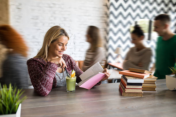 Young happy woman relaxing with a juice and book in a cafe while people in blurred motion are behind her.