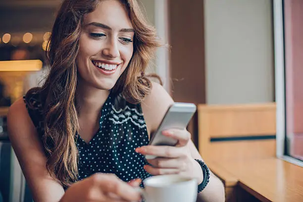 Smiling young woman holding coffee cup and dialing in cafeteria, with copy space