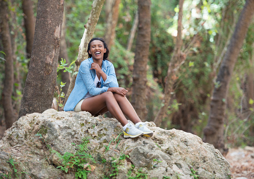 Happy smiling woman enjoying fresh air outdoors in forest, sitting on big rock stone,  looking at camera and laughing.