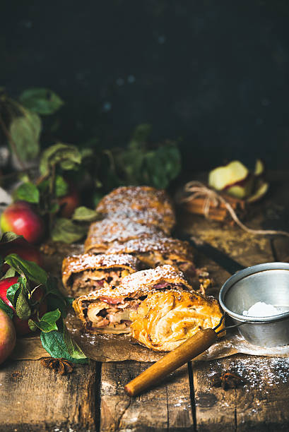 Apple strudel cake cut in pieces with fresh red apples Apple strudel cake cut in pieces with fresh red apples, sugar powder and spices on rustic wooden table, dark plywood wall background, copy space, vertical composition apple strudel stock pictures, royalty-free photos & images