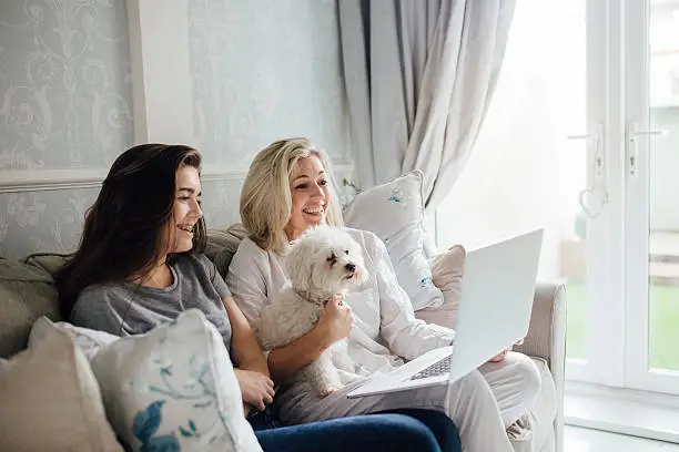 Mother and her daughter video calling someone on their laptop. They are smiling and waving at the screen. Their dog is sitting with them.
