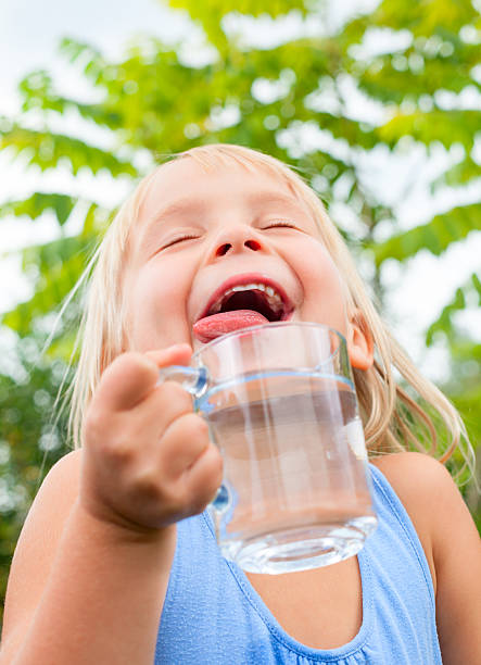 Blonde little girl wearing blue dress holding cup of fresh water with her eyes closed in a summer garden