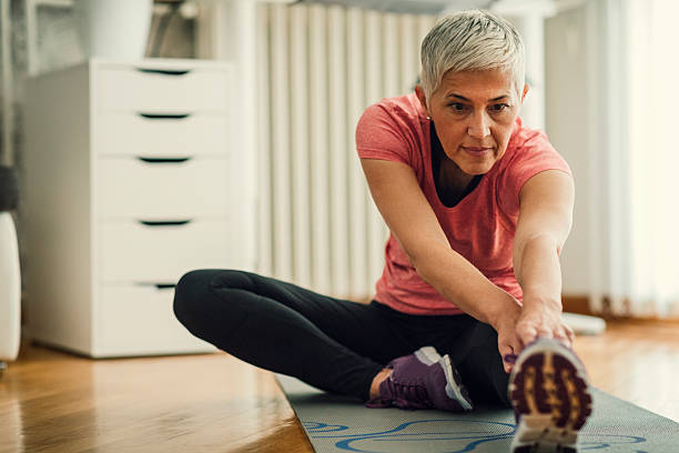 Mature Woman Exercise At Home. Mature woman exercising at home. Sitting on exercise mat and working stretching exercise , stretching her legs. stretched leg stock pictures, royalty-free photos & images