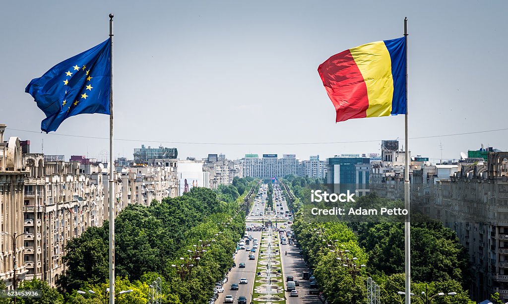 Romanian and European Union flag flying in Bucharest, Romania Close up image of the Romanian flag and the European Union (EU) flags flying at the top of the Palace of Parliament in Bucharest, Romania. In the distance we can see the main street of Bucharest stretching out into the distance, flanked by green trees and thronged with busy afternoon traffic. Horizontal colour image with copy space. Romania Stock Photo