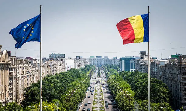 Close up image of the Romanian flag and the European Union (EU) flags flying at the top of the Palace of Parliament in Bucharest, Romania. In the distance we can see the main street of Bucharest stretching out into the distance, flanked by green trees and thronged with busy afternoon traffic. Horizontal colour image with copy space.