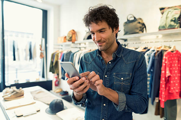 hombre enviando mensajes de texto en el teléfono celular en la tienda de ropa - market vendor fotos fotografías e imágenes de stock