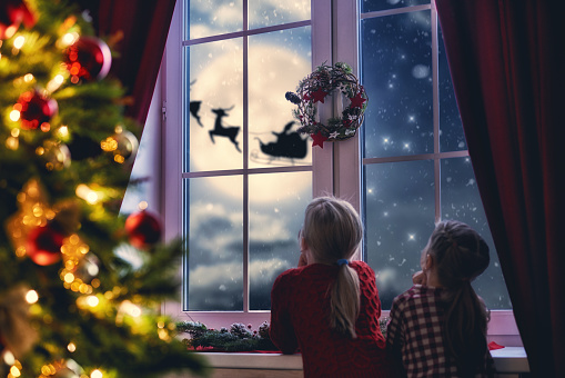 Merry Christmas and happy holidays! Cute little children girls sitting by window and looking at Santa Claus flying in his sleigh against moon sky. Room decorated on Christmas. Kids enjoy the holiday.