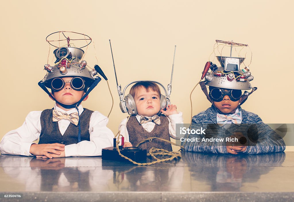 Three Boys Dressed as Nerds with Mind Reading Helmets A young boy imagines reading minds of his two friends with a homemade science project. They are dressed in casual clothing, glasses and bow ties. They are serious and sitting at a table with helmets on their heads in front of a beige background. Retro styling. Mental Health Professional Stock Photo