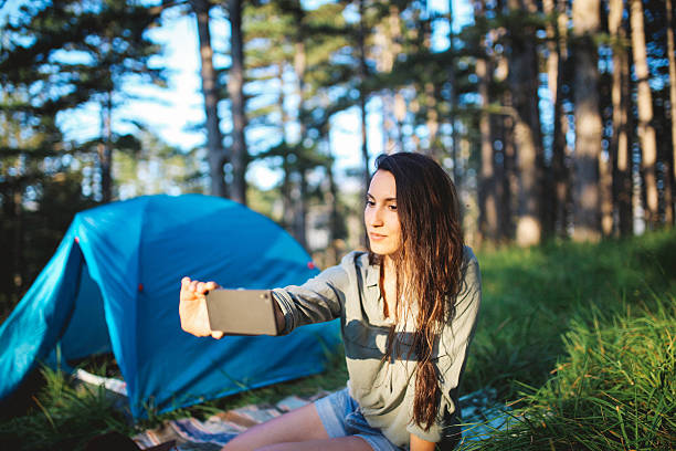 Young woman camping in the forest Beautiful young woman in vintage boho clothing camping in the forest in the Dinaric Alps in Southeastern Europe, on the border of Bosnia and Croatia. She is enjoying a nice early morning in the nature, having a warm cup of coffee, relaxing in the grass, taking a selfie with a smartphone. women lying down grass wood stock pictures, royalty-free photos & images