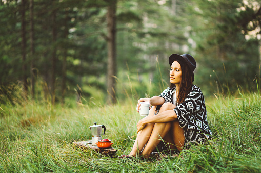 Beautiful young woman in vintage boho clothing camping in the forest in the Dinaric Alps in Southeastern Europe, on the border of Bosnia and Croatia. She is enjoying a nice early morning in the nature, having a warm cup of coffee, relaxing in the grass.