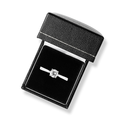 A sparkling solitaire diamond ring is in black leather ring box on a velvet cushion. The 1-carat diamond is set on a platinum band. The ring box is on a white background with a clipping path. Shot from overhead, looking down into the box.