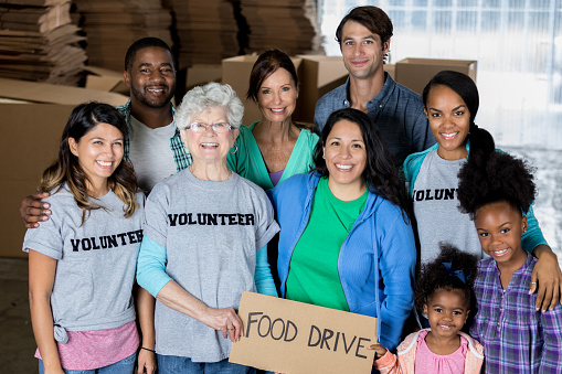 Caucasian, Hispanic and African American volunteers pose for a group photo while working at a food bank. They are holding a 'Food Drive' sign.