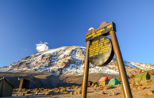 Kilimanjaro, Tanzania - March 9, 2015: Kibo with Uhuru Peak (5895m amsl) at Mount Kilimanjaro, Kilimanjaro National Park, seen from Karanga Camp at 3995m amsl. Park sign and tents in the background.