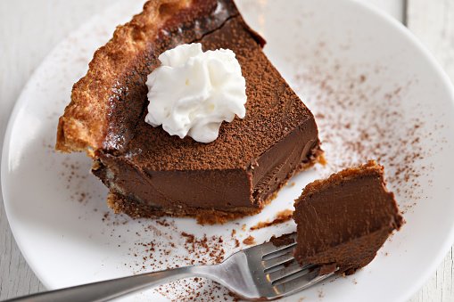 An extreme close up overhead view of a slice of chocolate Derby pie, dusted with powdered cocoa and topped with white whipped cream, a fork with a piece of pie on it lays on the plate.