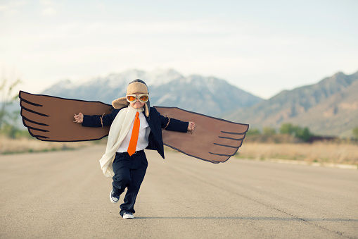 A young business boy dressed in suit, tie and flying goggles runs with his cardboard bird wings to the sky. He is dreaming of flying and becoming a pilot. His business is flying. Image taken in Utah, USA.