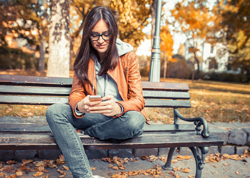 Portrait of young urban style woman sitting on bench at park and typing message on smart phone.Autumn season.