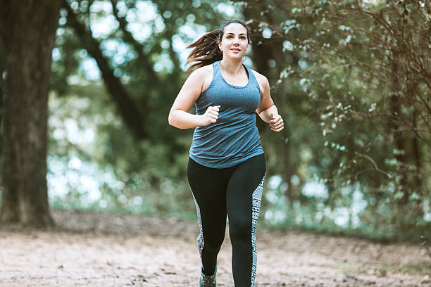 Woman Running in Park A young adult woman goes for a morning run, jogging on the park paths along the Colorado river by downtown Austin, Texas.  She has a content smile on her face.  Horizontal image. body confidence stock pictures, royalty-free photos & images