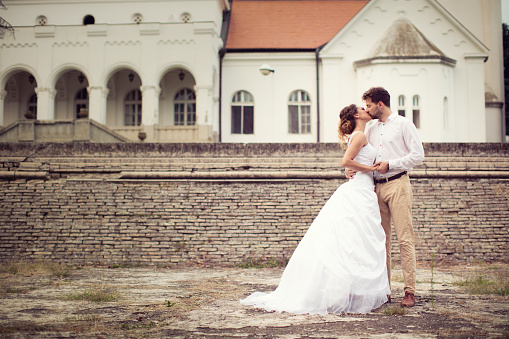 Just married couple kissing each other in front of old castle. She has a beautiful wedding dress with big veil and he is wearing elegant suit