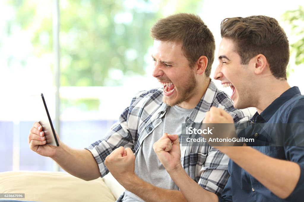 Excited friends watching tv on a tablet Two excited friends or roommates watching tv on line in a tablet sitting on a couch in the living room at home Sport Stock Photo