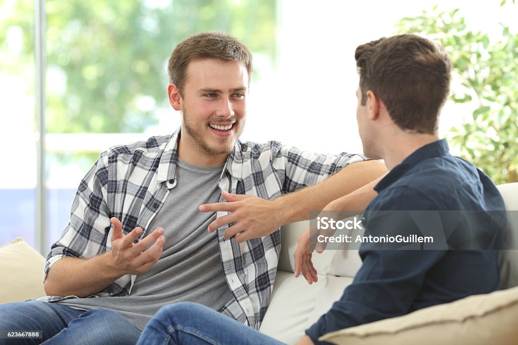 Two friends talking at home Two friends talking sitting in a couch in the living room with a window in the background at home Discussion Stock Photo