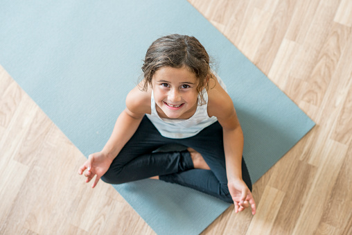 An elementary age girl is meditating and doing yoga on an exercise mat. She is smiling and looking up at the camera.