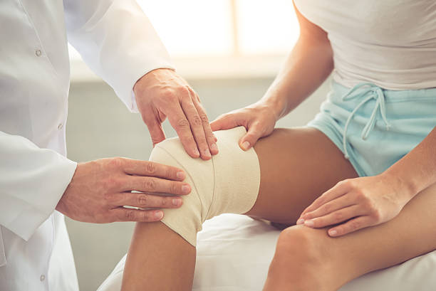 Doctor and patient Cropped image of handsome doctor bandaging woman's injured knee while working in his office physical therapy recovery touching human knee stock pictures, royalty-free photos & images