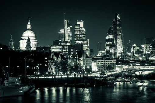 Night time image displaying the famous London skyline illuminated at night in all its glory. In foreground the silky surface of the river Thames, with city lights reflected in the still waters. On the skyline we can see such famous sights as St Pauls Cathedral and the offices of The Shard, the tallest building in western Europe, aglow with light. Horizontal monochrome toned image with copy space.
