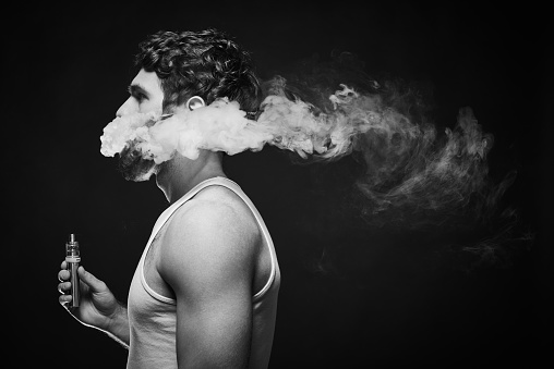 Bearded man with stylish haircut in profile holding electronic cigarette in hand and exhaling smoke in air, black and white waist-up shot