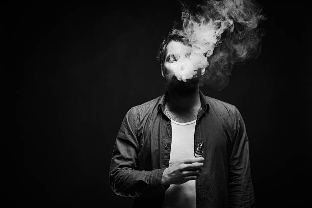 Brutal vaper in denim shirt Waist-up portrait of bearded man in denim shirt holding electronic cigarette in hand, blowing out smoke cloud and looking at it against black background, black and white shot black and white men facial hair beard stock pictures, royalty-free photos & images