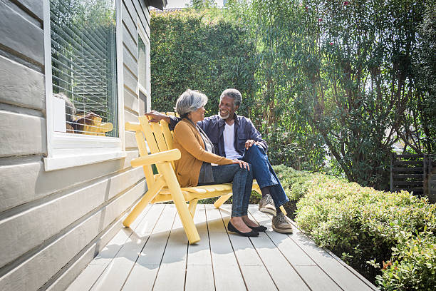 Senior African American couple sitting on porch outside house They are sitting on a yellow bench on the decking together outdoors in the garden porch stock pictures, royalty-free photos & images