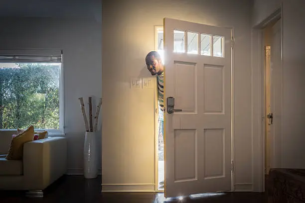 Young man peering into house through crack in from door with sunlight behind