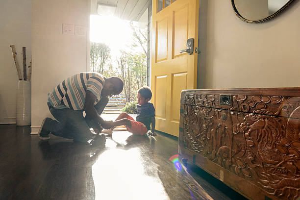 African American father helping son with shoes by front door The young boy is sitting on the floor in sunlight as his father helps him put his shoe on child candid indoors lifestyles stock pictures, royalty-free photos & images