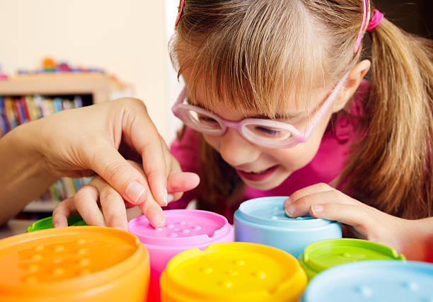 Smiling child with disability touching textured cups with her teacher Little girl with poor vision is playing with colorful tactile toy cups as a part of occupational therapy with her teacher. occupational therapy photos stock pictures, royalty-free photos & images