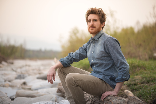 Young hipster man sitting on rocks next to a river and looking away, nature and healthy lifestyle concept